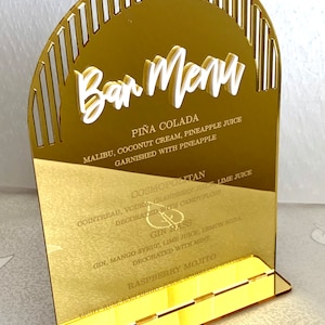 May include: Gold acrylic bar menu sign with a white arch and white text listing cocktails: Pi&ntilde;a Colada, Cosmopolitan, Gin Hass, and Raspberry Mojito.