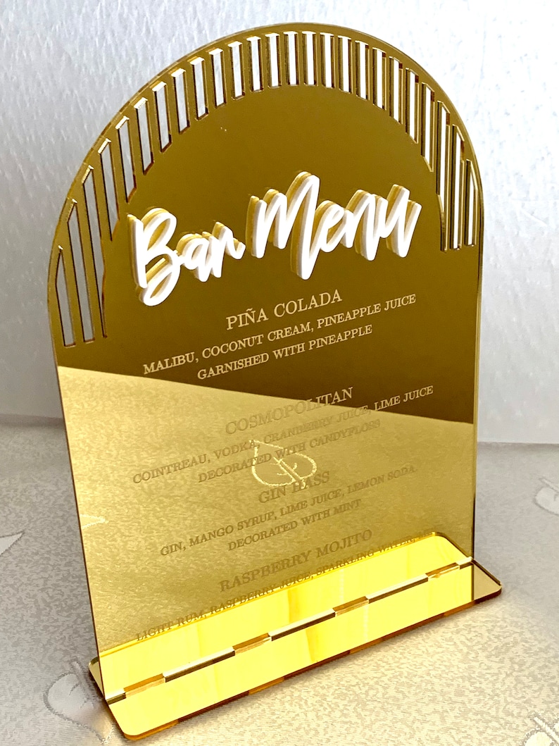 May include: Gold acrylic bar menu sign with a white arch and white text listing cocktails: Pi&ntilde;a Colada, Cosmopolitan, Gin Hass, and Raspberry Mojito.