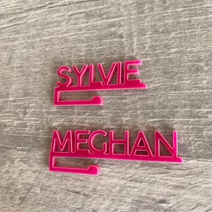 Two pink plastic nameplate holders with the names "SYLVIE" and "MEGHAN" printed in white.