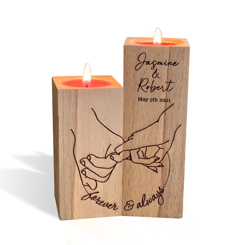 May include: Two wooden candle holders with a hand-drawn design of two hands intertwined. The candle holders are engraved with the text "Jasmine & Robert May 5th 2021 Forever & Always".
