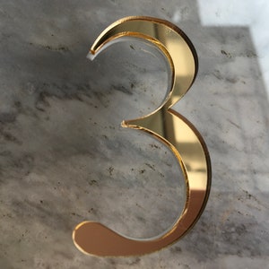 May include: A gold-colored number three, made of reflective material, is displayed on a gray marble surface.