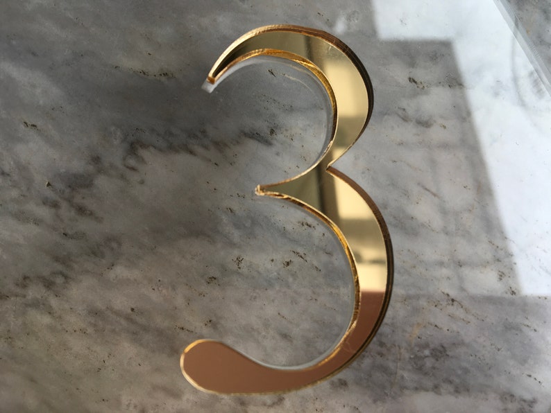 May include: A gold-colored number three, made of reflective material, is displayed on a gray marble surface.