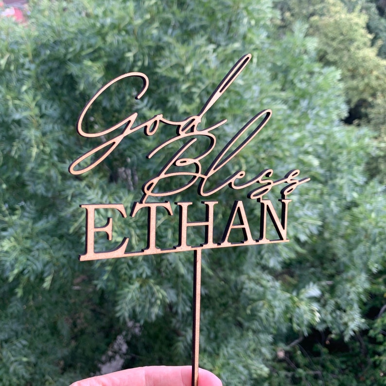 以下が含まれることがあります： 「God Bless Ethan」というテキストが書かれた木製のケーキトッパー。トッパーは茶色で、ケーキに差し込むための棒がついています。
