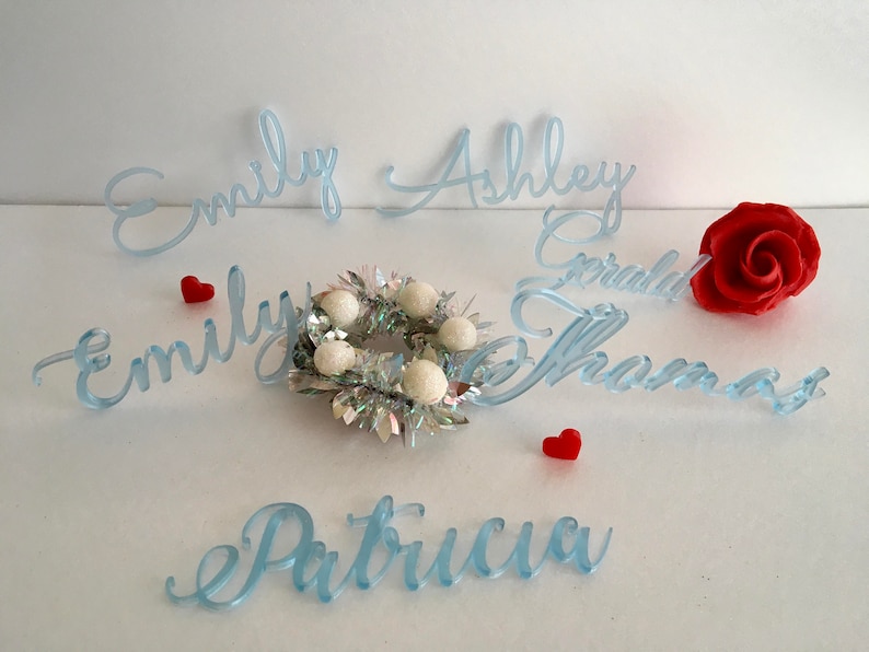 Peut inclure: D&eacute;coupes de pr&eacute;noms en acrylique bleu clair avec les noms Emily, Ashley, Gerald, Thomas et Patricia. Une petite couronne en guirlande argent&eacute;e avec des pompons blancs et deux petits c&oelig;urs rouges accompagnent une rose rouge.