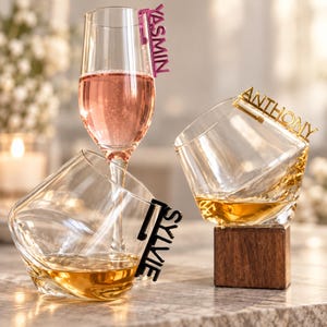 Peut inclure: Une flûte à champagne remplie de liquide rose avec une étiquette nominative en acrylique rose YASMIN, et deux verres à whisky inclinés contenant du liquide ambré avec des étiquettes en acrylique noir et or SYLVIE et ANTHONY.