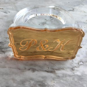 May include: A clear acrylic napkin ring with a gold-colored metal plate featuring the initials "P&K" in a decorative script font.