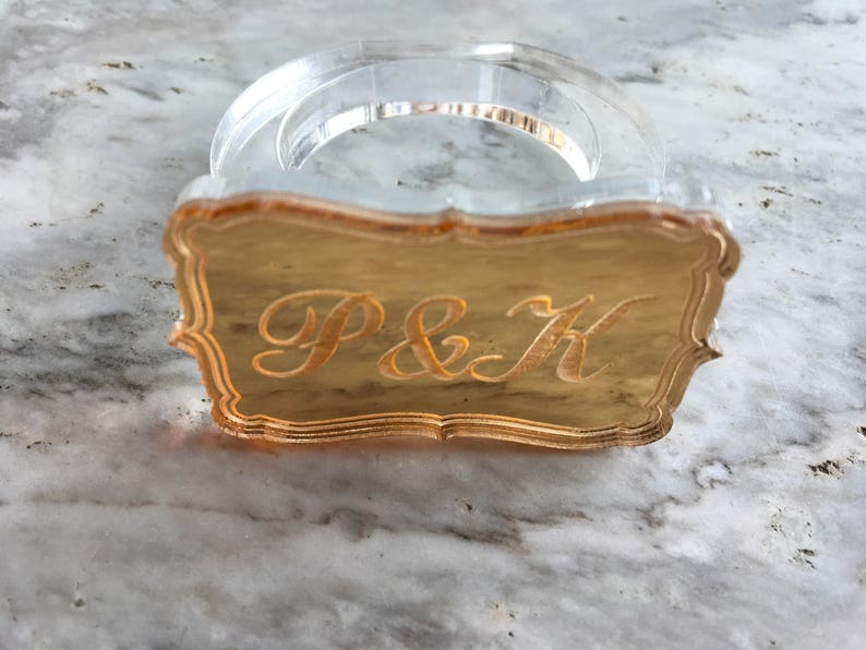 May include: A clear acrylic napkin ring with a gold-colored metal plate featuring the initials "P&K" in a decorative script font.