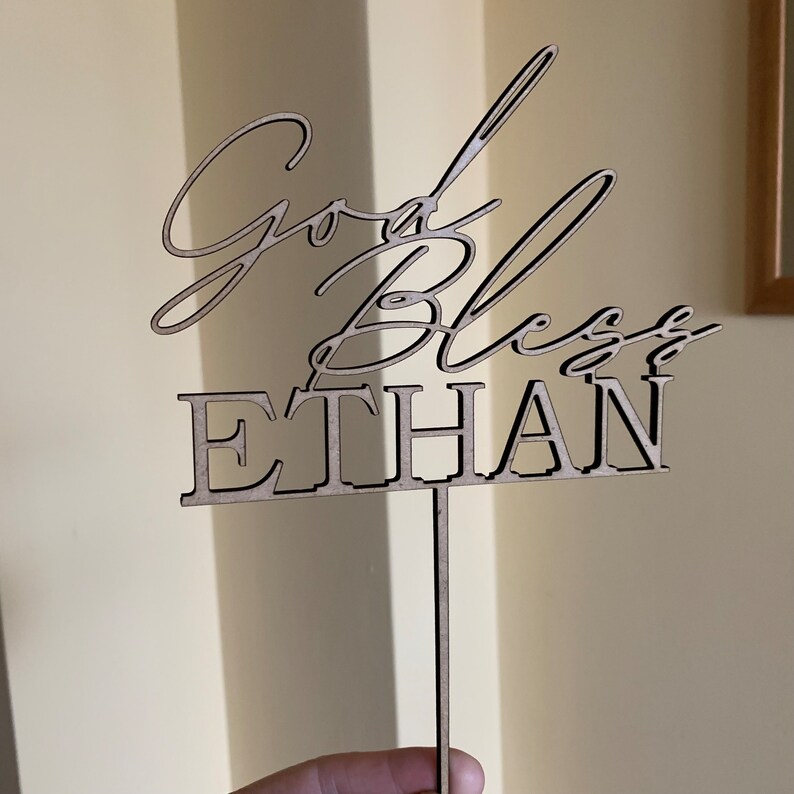 以下が含まれることがあります： 「God Bless Ethan」というテキストが書かれた木製のケーキトッパー。トッパーは茶色で、ケーキに差し込むための棒がついています。