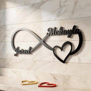 Può includere: Segno infinito in metallo nero con i nomi "Jared" e "Mckayla" in corsivo, un cuore e due oggetti a forma di cuore dorati e rossi. L'insegna è montata su una parete di marmo bianco e grigio.