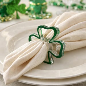 Op de afbeelding: Een groene klavervormige servetring houdt een crèmekleurige linnen servet op een wit bord. De servetring heeft een doorzichtig midden en een groene omtrek. Op de achtergrond zijn andere St. Patrick's Day-decoraties te zien.