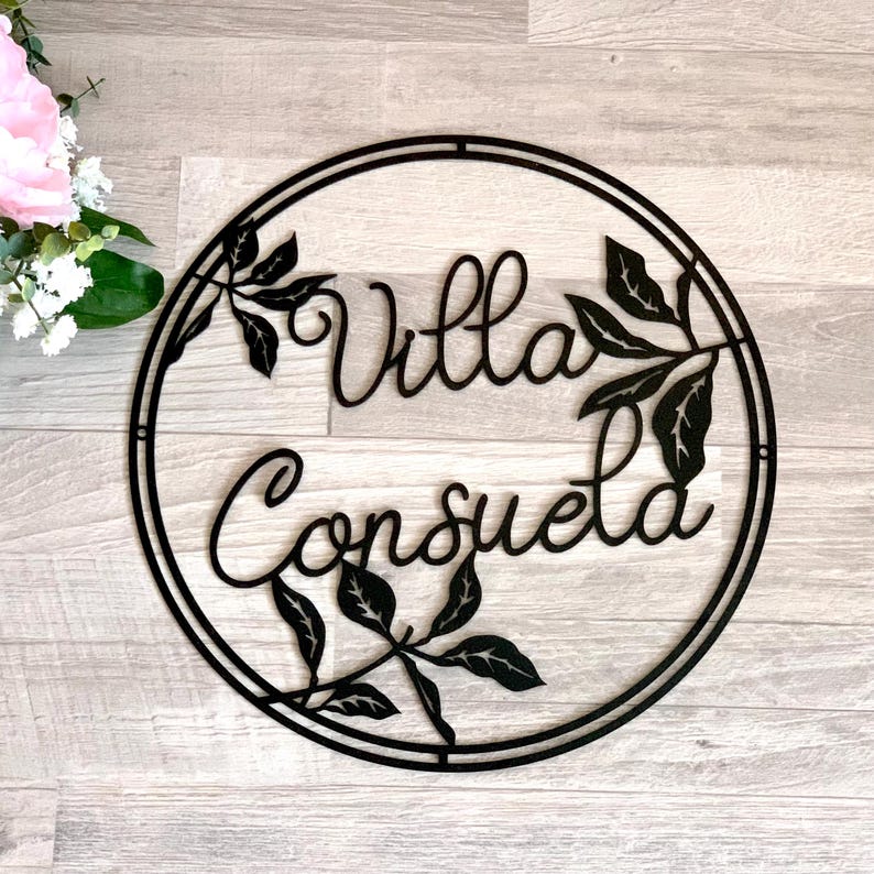 May include: Black metal sign with a circular frame and the words "Villa Consuela" in a decorative script font. The sign is decorated with stylized leaves.