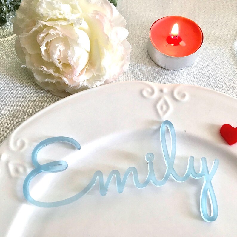 Peut inclure: Une plaque nominative en acrylique bleu clair portant le pr&eacute;nom Emily &eacute;crit en lettres cursives, pos&eacute;e sur une assiette en c&eacute;ramique blanche avec des d&eacute;tails d&eacute;coratifs en relief sur le bord.