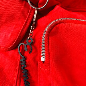 May include: A red backpack with a black metal zipper and a black metal keychain with the word "Emma" written on it.