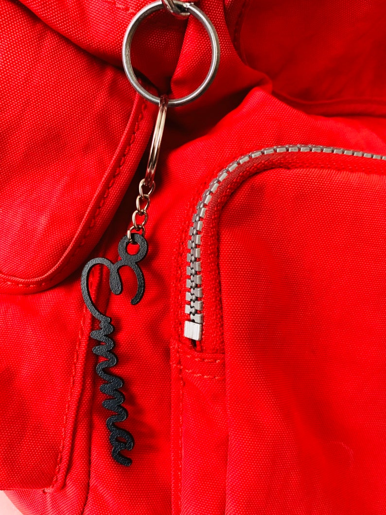 May include: A red backpack with a black metal zipper and a black metal keychain with the word "Emma" written on it.