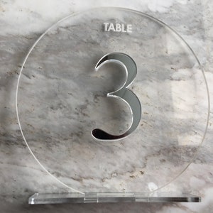 May include: A clear acrylic table number with the word "TABLE" and the number "3" in a silver reflective finish.