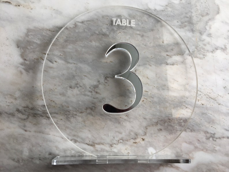 May include: A clear acrylic table number with the word "TABLE" and the number "3" in a silver reflective finish.