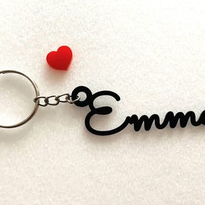 May include: Black keychain with the name "Emma" in a cursive font. The keychain has a silver ring and a small red heart charm.