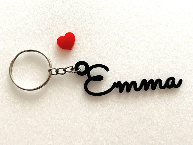 May include: Black keychain with the name "Emma" in a cursive font. The keychain has a silver ring and a small red heart charm.