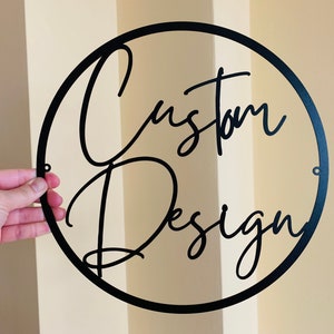 Pode incluir: Uma placa de metal preta com as palavras "Custom Design" em fonte cursiva, dentro de uma moldura circular. A placa tem pequenos orifícios para pendurar e é segurada por uma mão contra um fundo neutro.