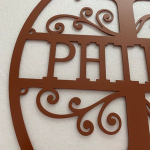 May include: A brown metal family name sign with the names "JOHN" and "PHILIP" cut out. The sign features a tree design with decorative scrollwork and a circular frame. The metal has a matte finish.
