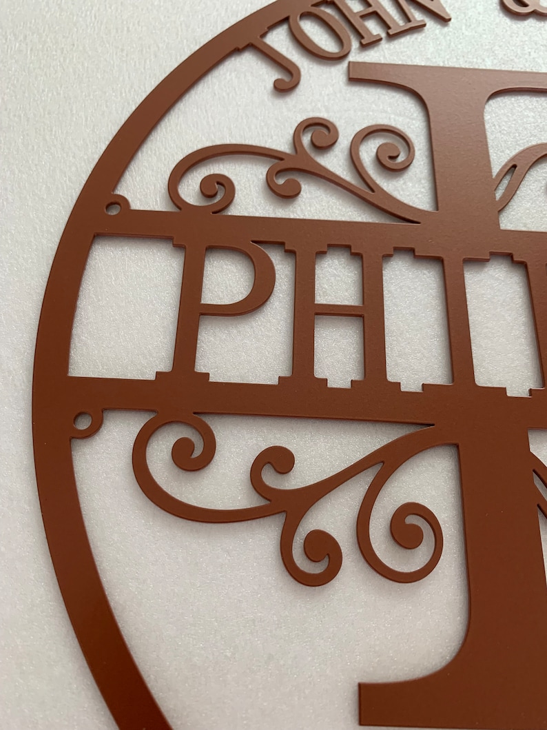 May include: A brown metal family name sign with the names "JOHN" and "PHILIP" cut out. The sign features a tree design with decorative scrollwork and a circular frame. The metal has a matte finish.