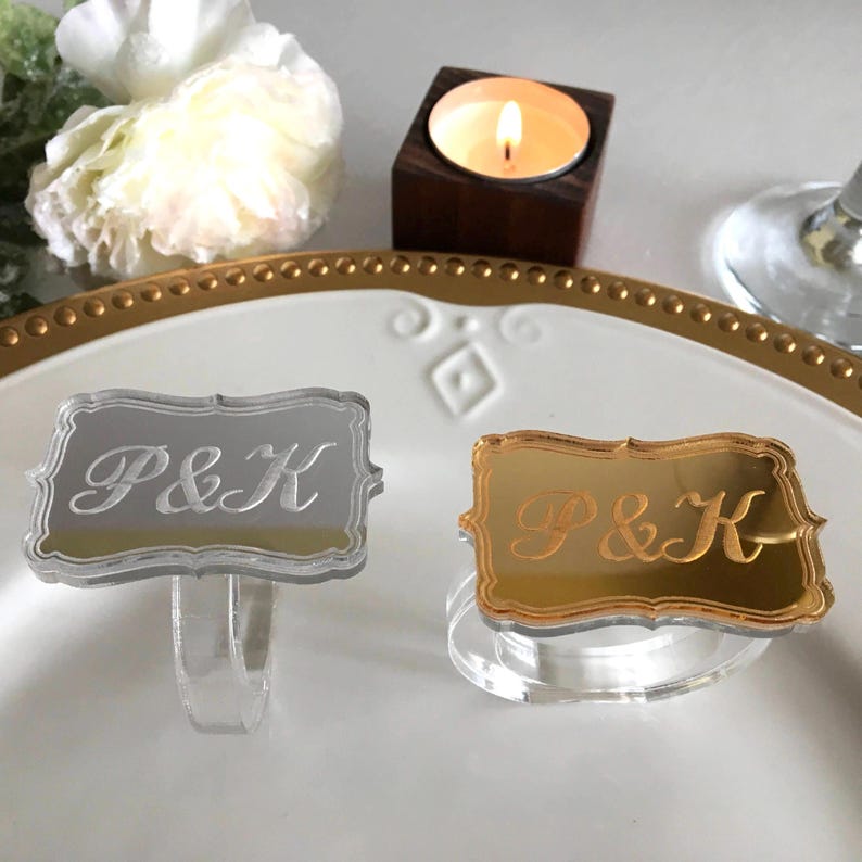 May include: Two acrylic napkin rings with mirrored surfaces. One ring is silver with white engraved lettering that says "P&K". The other ring is gold with gold engraved lettering that says "P&K".