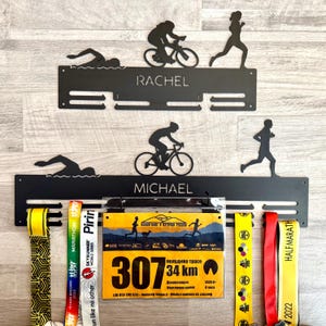 May include: Black metal medal display rack with cut-out silhouettes of a swimmer, cyclist, and runner. The rack has slots for medals and the names "RACHEL" and "MICHAEL". Includes race bibs and colorful ribbons.