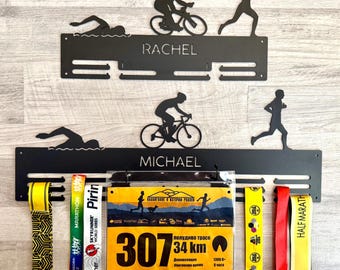 Könnte beinhalten: Schwarzes Metall-Medaillen-Display mit ausgeschnittenen Silhouetten eines Schwimmers, Radfahrers und Läufers. Das Rack hat Schlitze für Medaillen und die Namen "RACHEL" und "MICHAEL". Enthält Startnummern und bunte Bänder.