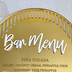 May include: Gold mirror bar menu sign with a white, cursive "Bar Menu" title. The sign lists a "Pi&ntilde;a Colada" cocktail recipe with Malibu, coconut cream, pineapple juice, and a pineapple garnish.
