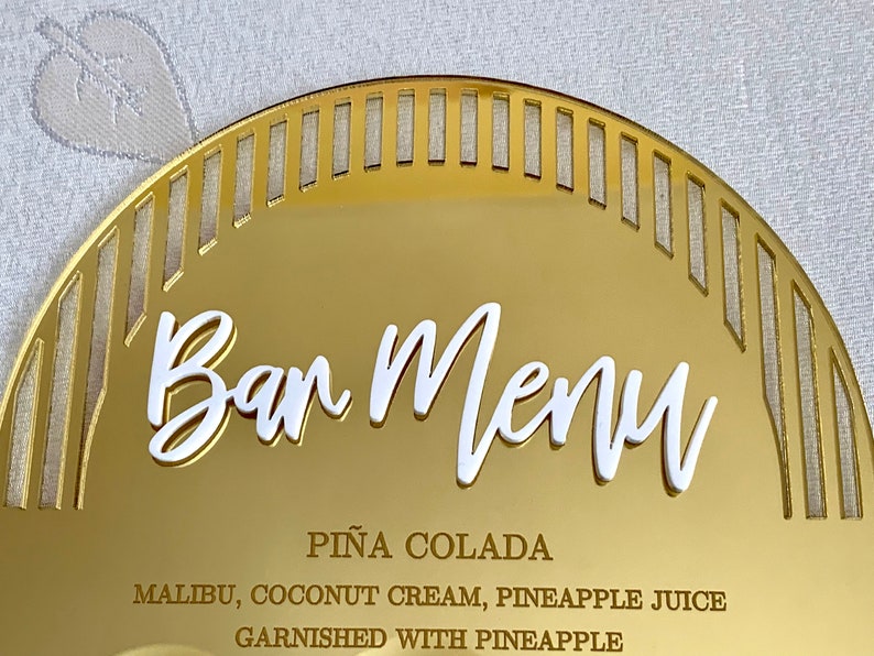 May include: Gold mirror bar menu sign with a white, cursive "Bar Menu" title. The sign lists a "Pi&ntilde;a Colada" cocktail recipe with Malibu, coconut cream, pineapple juice, and a pineapple garnish.