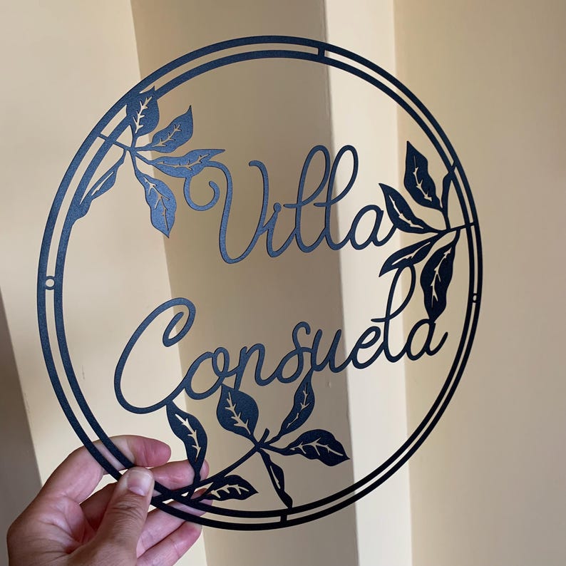 May include: Black metal circular sign with the words "Villa Consuelo" in a decorative script font. The sign is decorated with cut-out leaf designs.