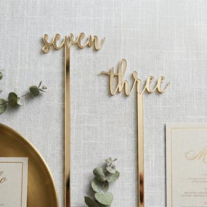 Pode incluir: Decorações douradas para números de mesa com os números "seven" e "three" em fonte cursiva. Os números estão presos a hastes longas e finas de ouro. O fundo é uma superfície com textura de linho cinza claro.