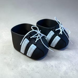 May include: A pair of miniature black and white trainers with white laces. The shoes have three white stripes on the sides, and are designed to look like athletic footwear. The shoes are likely for a doll or small child.