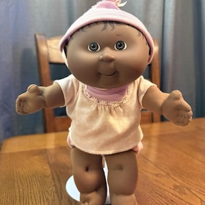 Puede incluir: Una muñeca bebé vintage con piel oscura, vestida con un atuendo rosa claro y un sombrero a juego. La muñeca tiene una expresión sonriente y está de pie sobre una base blanca. Los brazos de la muñeca están extendidos.