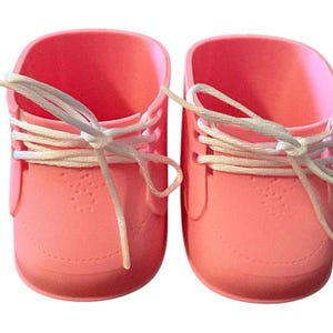 May include: A pair of coral-coloured baby shoes with white laces. The shoes have a rounded toe and a textured surface. The laces are tied in a bow. The shoes are made of a soft material.