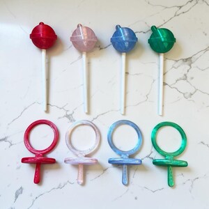 Op de afbeelding: Een assortiment kleurrijke lolly's en objecten in de vorm van een vrouwelijk symbool. De lolly's zijn rood, roze, blauw en groen, elk met een witte stok. De vrouwelijke symbolen zijn in bijpassende kleuren, met een cirkelvormige bovenkant en een kruisvormige basis.