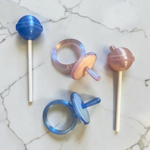 May include: Two spherical lollipops on white sticks and two ring-shaped toys with attached circular discs. One lollipop and one ring toy are blue, while the other lollipop and ring toy are a pale, shimmering pinkish-peach color.