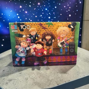 May include: A vintage MB 100 puzzle depicting a band of Cabbage Patch Kids dolls on a stage. The dolls are dressed in vibrant outfits and playing instruments, including drums, a guitar, and a microphone. The backdrop is a starry night scene.