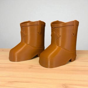 May include: A pair of brown cowboy boots. The boots are made of a solid material and have a classic western design, including decorative stitching and a pull tab. The boots are displayed on a wooden surface.