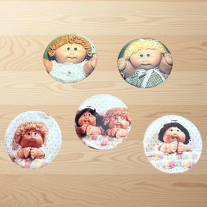 Puede incluir: Colección de cinco botones redondos con Cabbage Patch Kids. Los botones muestran varias muñecas con diferentes colores de pelo y atuendos. Los botones están colocados sobre un fondo de madera marrón claro.