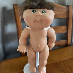 Może przedstawiać: Zabytkowa lalka Cabbage Patch Kid z brązowymi włosami i zielonymi oczami. Lalka ma opaloną karnację i stoi na białym stojaku. Lalka ma na sobie biały naszyjnik.