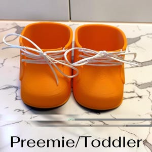 May include: A pair of bright orange shoes with white laces, featuring a rounded toe design and a smooth finish. The text "Preemie/Toddler Size" is printed in black below the shoes, indicating the intended scale for these footwear items.