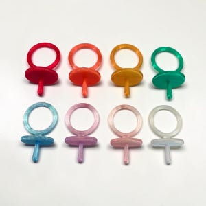May include: Eight colorful ring-shaped bottle stoppers in red, orange, yellow, green, blue, purple, pink, and white. Each stopper has a circular ring and a flat, round base with a stem.