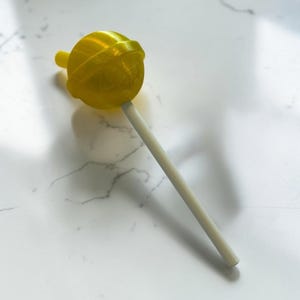 Könnte beinhalten: Ein gelber Lutscher mit einem runden Bonbonkopf und einem weißen Holzstab. Das Bonbon hat eine strukturierte Oberfläche und ist diagonal vor einem weißen Marmorhintergrund positioniert.