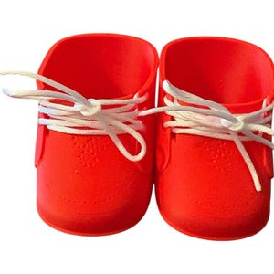 May include: A pair of vibrant red baby shoes with white laces. The shoes feature a textured surface and a classic sneaker design, suitable for a newborn or infant. The laces are tied in a bow.