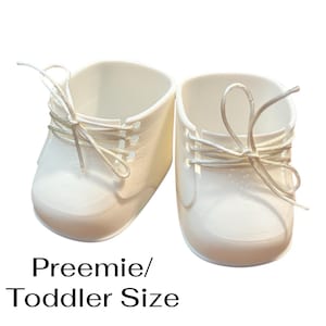 Może przedstawiać: Para białych bucików dla niemowląt w stylu oksfordzkim, ze sznurowadłami. Buty wykonane są z gładkiego materiału i przeznaczone są dla wcześniaków i małych dzieci. Poniżej butów widnieje napis "Preemie/Toddler Size".