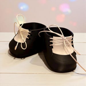 May include: A pair of black and white doll shoes with cream-colored laces. The shoes have a classic design with a black toe and heel, and a white section on the top. The laces are tied in a bow.