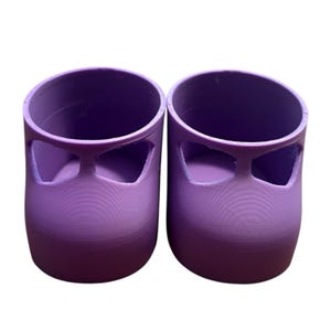 May include: Two matching purple 3D-printed objects, resembling small shoes or containers. Each has a cylindrical body with a rounded top and a rectangular cutout near the top. The objects are made of a solid, smooth material.