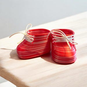May include: A pair of red and white striped baby booties with white laces. The shoes are made of a translucent material and sit on a light-colored wooden surface. The booties are designed to look like miniature sneakers.