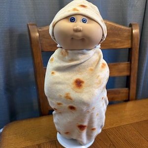 Op de afbeelding: Een Cabbage Patch Kid-pop gewikkeld in een tortilla-deken. De pop heeft blauwe ogen en een neutrale uitdrukking. De tortilladeken heeft bruine vlekken en bedekt het lichaam en het hoofd van de pop.